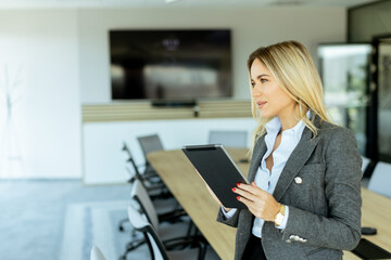 Obraz premium Smiling professional woman in red blazer holding tablet in modern conference room during daytime
