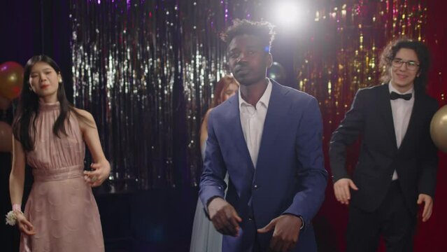 Youthful African American guy in classy suit standing in front of his classmates in evening attire while dancing at disco in the night club during prom celebration