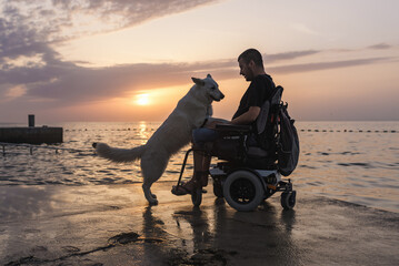 Man with disability, electric wheelchair user petting his dog and enjoying a sunset on the beach....
