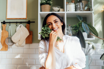 Attractive young woman stands in the kitchen and holds a bunch of fresh mint. A charming brunette...