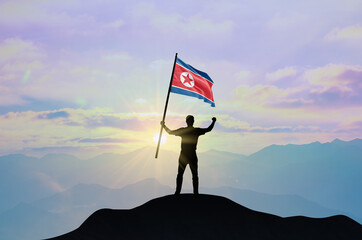 Obraz premium North Korea flag being waved by a man celebrating success at the top of a mountain against sunset or sunrise. North Korea flag for Independence Day.