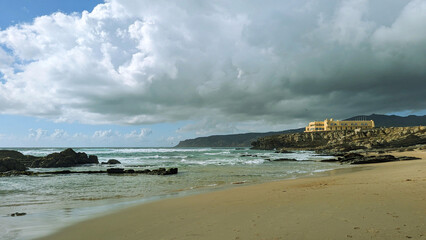 Scenic View of a Sandy Beach with a Building on a Cliff and Dramatic Clouds Over the Ocean