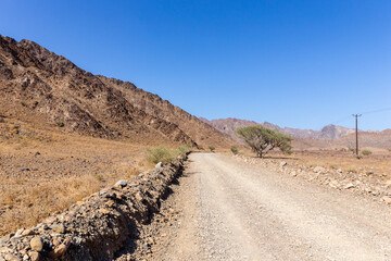 Gravel dirt road through rocky limestone Hajar Mountains and cliffs in United Arab Emirates, barren desert vegetation and electrical pylons along the road, desert landscape.