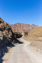 Winding gravel dirt road through rocky limestone Hajar Mountains in United Arab Emirates, electrical pylons along the road.