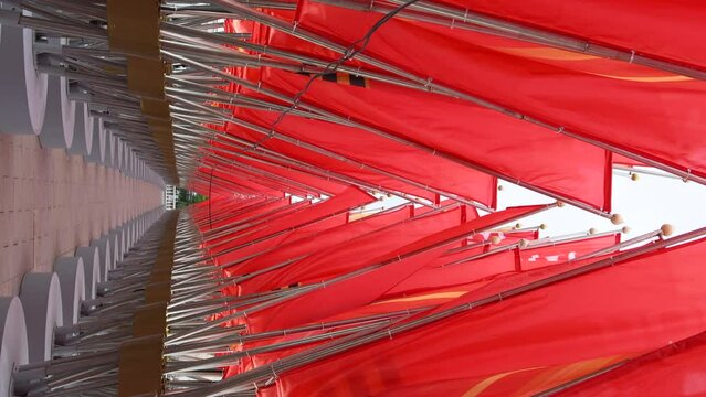 Close-up side view of empty arch decoration made from swaying red flags or banners on city square. Soft focus. Real time handheld vertical video. National holidays decorations theme.