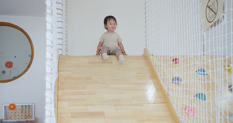 A young child is sitting on a wooden slide and play slider down with fun. The slide is located in a...