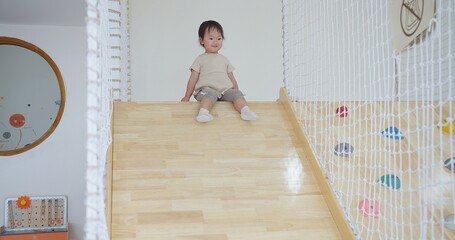 A young child is sitting on a wooden slide and play slider down with fun. The slide is located in a...