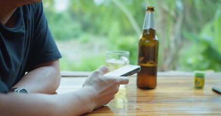 A Asian man is sitting at a table using a cell phone in his hand and a bottle of beer next to him