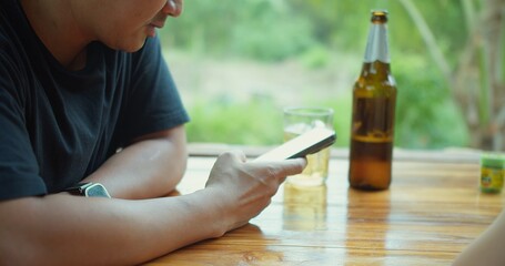 A Asian man is sitting at a table using a cell phone in his hand and a bottle of beer next to him