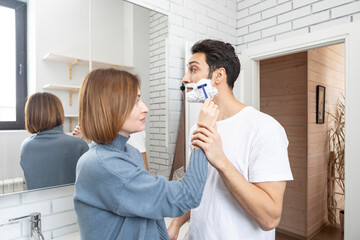 Young attractive woman helps her husband shaving in the bathroom