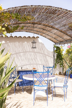Table and chairs underneath a palapa with sunlight streaming through and striking shadows in a garden in Algarve, Portugal. 