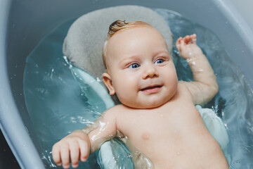 A small blue-eyed baby bathes in a bath on a stand. A baby is bathing and smiling.