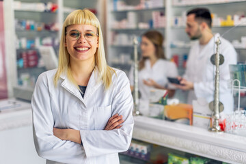 Smiling portrait of young, beautiful female pharmacist that is standing at the cash desk. Other...