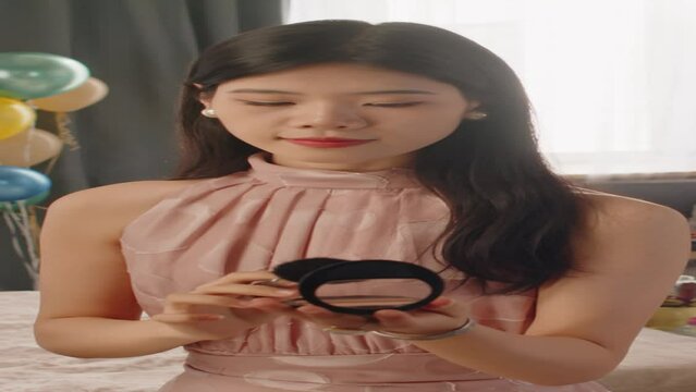 Vertical shot of Asian teenage girl in pink evening gown taking a bit of compact powder with cosmetic brush and applying it on her face while preparing for prom party
