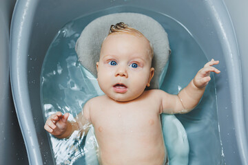 A small blue-eyed baby bathes in a bath on a stand. A baby is bathing and smiling.
