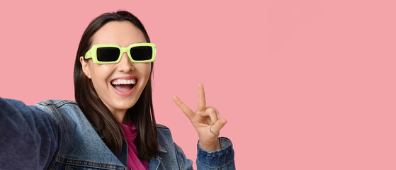 Beautiful young happy woman in stylish denim jacket and sunglasses taking selfie on pink background