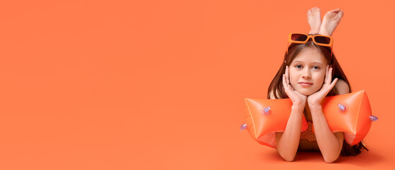 Cute little happy girl in swimsuit with inflatable armbands lying on orange background