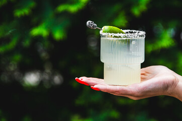 A hand holding a salt-rimmed glass with lime and a beverage against a blurred green background