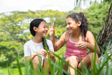 two brunette latina girls, sitting next to a tree in a park playing and laughing together