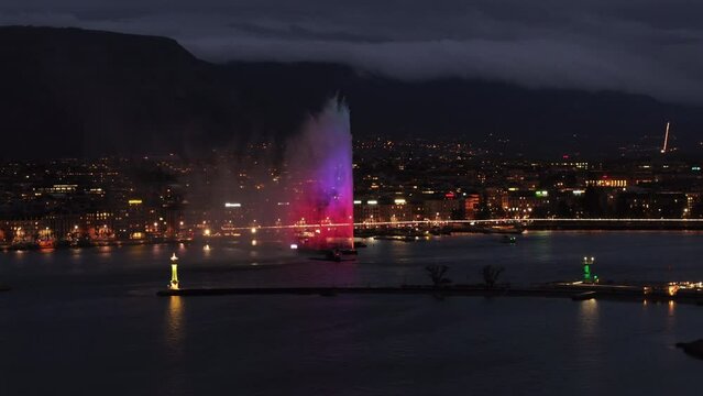 Geneva's jet d'eau fountain illuminated at night, casting vibrant colors against the cityscape and Lac Leman water