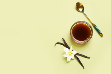 Vanilla extract in bowl with spoon, vanilla pods and flowers on green background. Top view