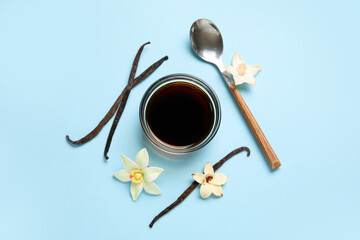 Vanilla extract in bowl with spoon, vanilla pods and flowers on blue background. Top view