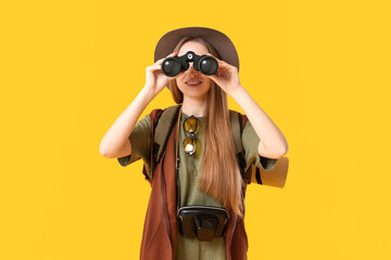 Smiling young woman looking through binoculars on yellow background. Travel concept