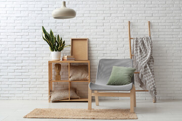 Armchair with cushion, shelving unit with plant and ladder with plaid near white brick wall
