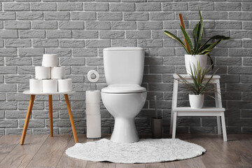 Interior of restroom with toilet bowl, coffee table with toilet paper and plants near grey brick...