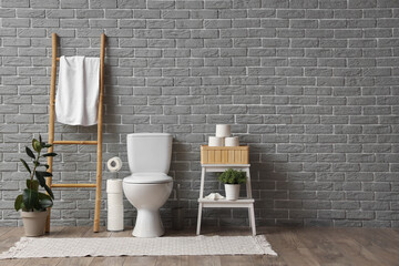 Interior of restroom with toilet bowl, ladder and shelving unit with toilet paper near grey brick...