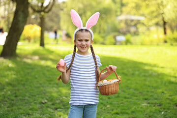 Easter celebration. Cute little girl in bunny ears holding wicker basket and painted egg outdoors
