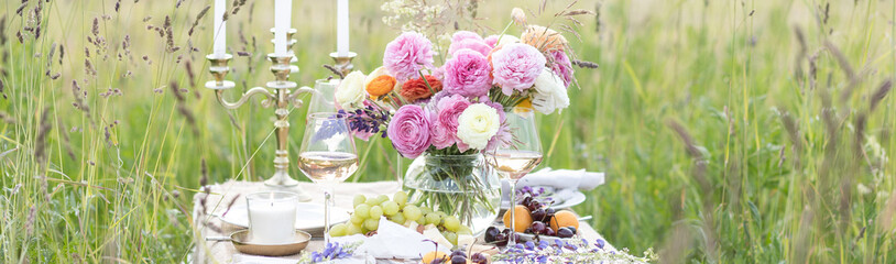 Elegant wedding or private party for a loving couple table setting outdoors in the garden. Beautiful decor with fresh ranunculi flowers, candles. Gourmet cheese board, fruits, white wine. Banner