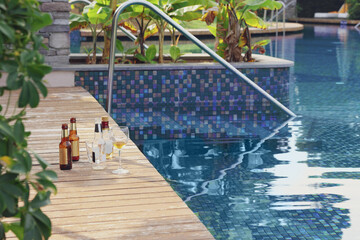 Wooden deck with bottles of drink and glasses near outdoor swimming pool. Luxury resort