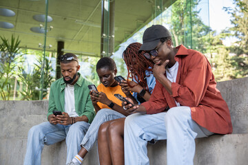 Four immersed in smartphones, African American friends sit together outside university library...