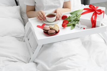 Tasty breakfast served in bed. Woman with tea, macarons, gift box, flowers and I Love You card at...