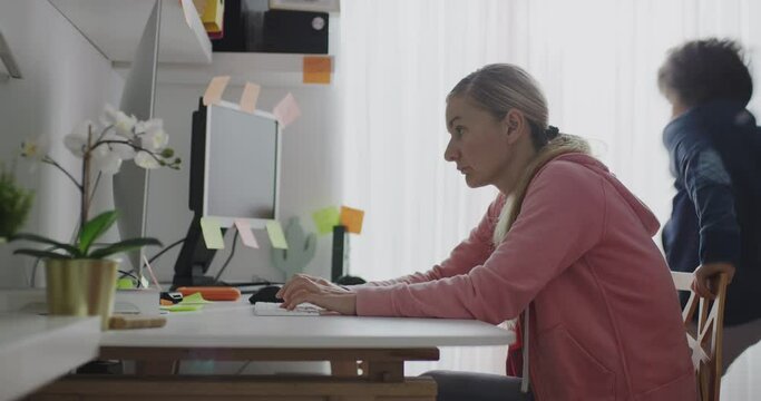 A woman attempts to work from home while her two children engage in distracting antics beside her at a desk.