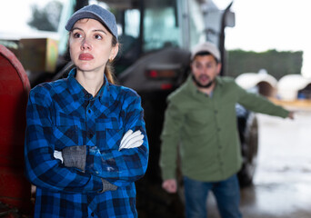 Upset confused young workwoman standing near tractor in farm backyard while angry farmer...