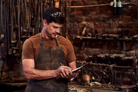 Metal worker, clipboard and writing notes in workshop for manufacturing, welding and blacksmith. Tradesman, tools and equipment in factory with apron for artisan, industrial steel and manual labor - Powered by Adobe