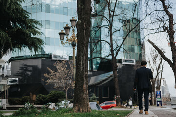 A suited businessman with a briefcase walks on a sidewalk with trees and street lamps, heading towards glass-fronted skyscrapers.