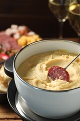 Dipping piece of sausage into fondue pot with melted cheese at table, closeup