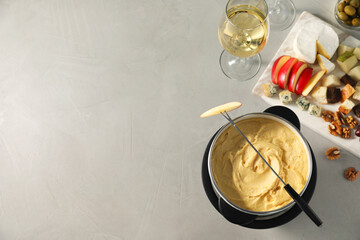 Fork with piece of apple, melted cheese in fondue pot and snacks on grey table, flat lay. Space for...