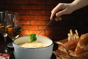 Woman dipping piece of broccoli into fondue pot with melted cheese at wooden table with wine and...