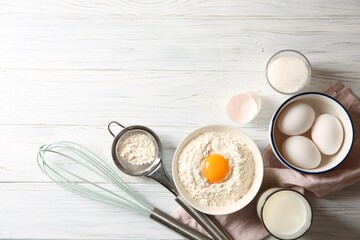 Flour with yolk in bowl and other ingredients for dough on white wooden table, flat lay. Space for...