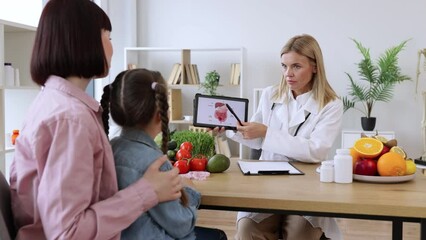 Mature experienced female doctor showing tablet with gastrointestinal tract image for treatment planning to little female child. Mother with little girl at appointment with family nutritionist.