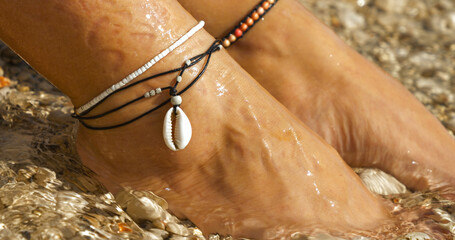 CLOSE UP: Elegant anklets with a seashell on female feet by the sparkling sea, reflecting a perfect...