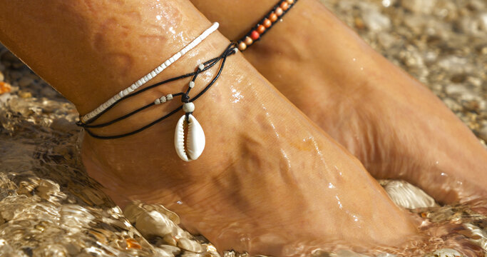 CLOSE UP: Elegant anklets with a seashell on female feet by the sparkling sea, reflecting a perfect blend of style and summertime serenity on a pebble seashore. Relaxing holidays in sunny Dalmatia.