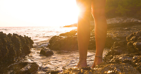 LENS FLARE, CLOSE UP: Lady admires golden sunset barefoot on a seashore where warm rays of setting...