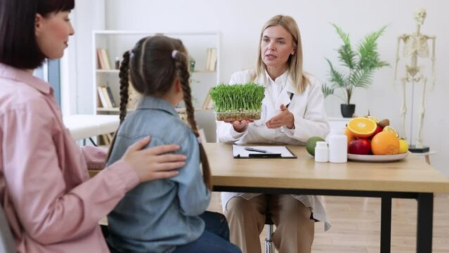 Beautiful female doctor holding microgreen while adding it to patients diet. Little girl sitting on mother lap at appointment with mature female nutritionist making healthy eating plan.