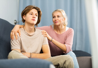 Friendly caring woman calming sad teenage son after disagreements, while sitting together in cozy living room..