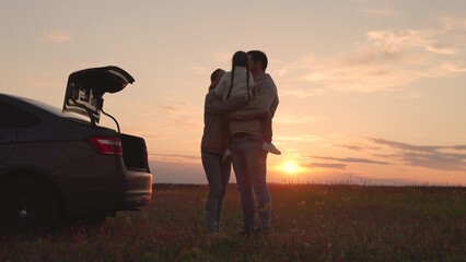 Dad, mother, child are hugging against backdrop of sunset next to car. Happy family traveling by...